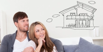 A couple sits on a couch smiling, envisioning their dream home depicted by a hand-drawn house sketch on the wall.