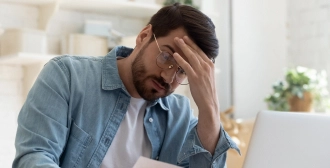 A man wearing glasses and a denim shirt holds his head in one hand, looking stressed as he reviews a document in a bright, home setting.