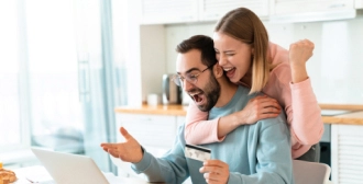 The image shows a man holding a credit card and looking thrilled at a laptop while a woman hugs him from behind and cheers, celebrating a happy moment in a kitchen.