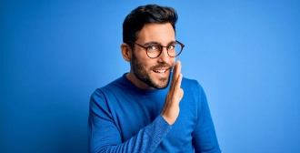 A man with glasses and a blue sweater leans forward and gestures as if whispering, set against a vibrant blue background.