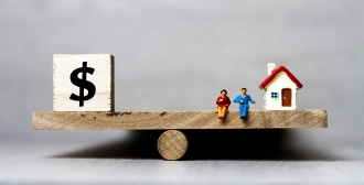 A wooden seesaw tilts under the weight of a dollar sign block on one side and a small house with two people on the other, representing financial considerations in real estate.