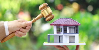 A gavel is held above a model house in an outdoor setting, representing a property auction or a significant decision in real estate, with greenery in the background.
