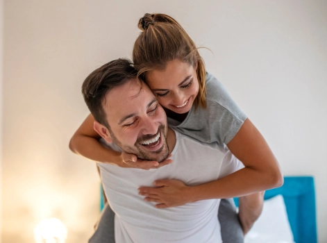 A cheerful couple shares a joyful moment, with the woman playfully riding on the man's back. They are both smiling and laughing, creating a warm and happy atmosphere in what appears to be their home.