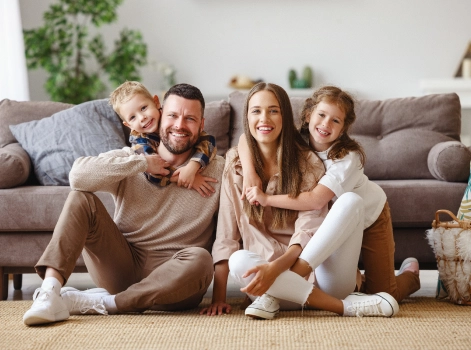 A happy family of four sitting on the floor in their living room. The parents are smiling, with their two young children, a boy and a girl, playfully hugging them. The scene exudes warmth and togetherness in a comfortable home setting.