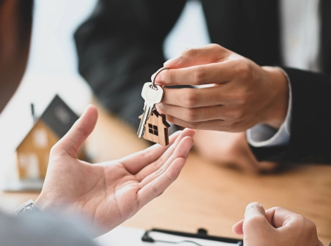 A close-up of a real estate agent handing over house keys to a new homeowner. The scene symbolises the completion of a property transaction, with a small house model visible in the background, highlighting the context of a home purchase.