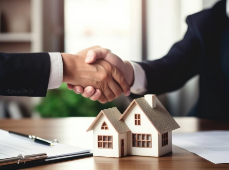 Two business professionals shaking hands over a desk, with a miniature house model and documents in the foreground. This image represents a successful real estate deal or mortgage agreement.