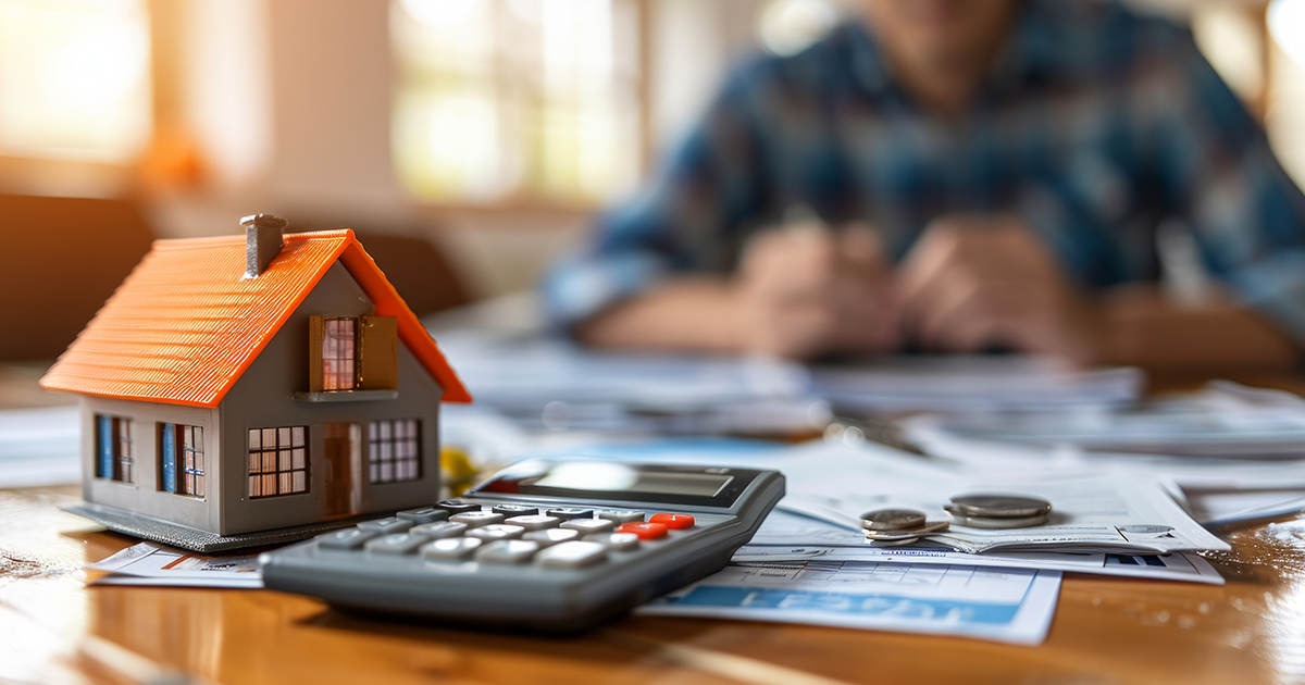 A miniature house and a calculator are placed on top of financial documents and coins, while a person works in the background. The scene hints at using a First Home Grant Calculator for home-buying decisions.