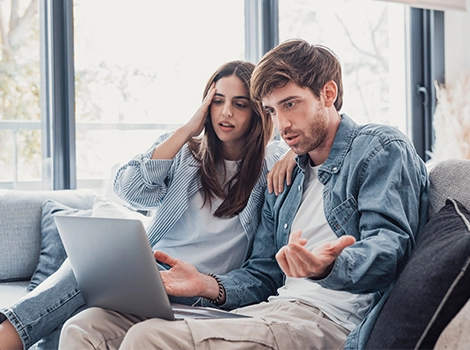 A young man gestures in frustration as his partner looks stressed, both seated by a laptop, reflecting home buyer’s remorse.