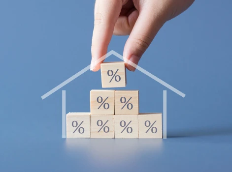 A blue background highlights a hand arranging percentage-marked wooden blocks into a pyramid, framed by a house outline, symbolizing refinancing home loans and interest adjustments.