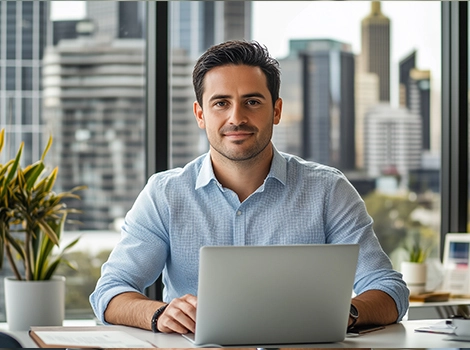 Confident man in a light blue shirt working on a laptop at a desk, with a cityscape visible through large office windows.