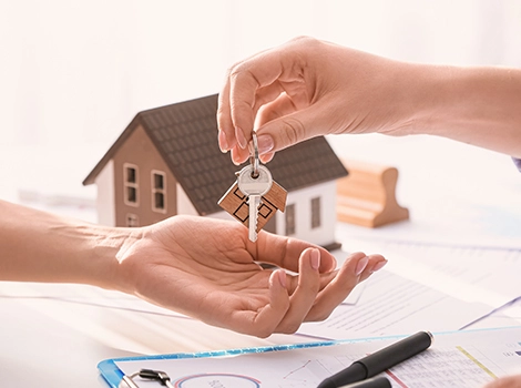 Hand giving house keys to another hand, with a model house and documents on the table in the background.