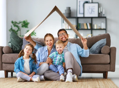 Happy family of four sitting together in their cozy living room, symbolizing security and comfort under a makeshift roof made of cardboard, representing the dream of homeownership through mortgage solutions