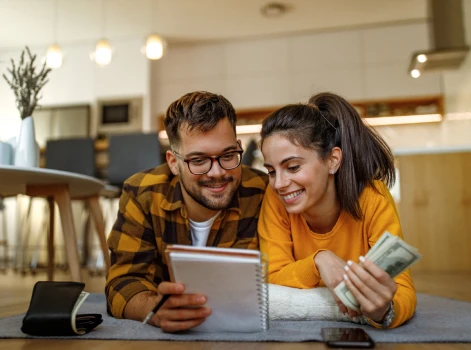 Young couple planning their finances at home, smiling as they review their notes and hold cash, symbolizing financial preparedness and the steps towards securing a mortgage.