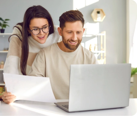 Smiling couple reviewing documents while using a laptop in a bright, modern home setting, happy after getting loan approval despite having bad credit and no deposit.