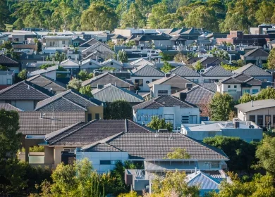 An aerial view of a suburban neighbourhood featuring a variety of modern houses with tiled roofs and lush greenery. The dense cluster of homes is surrounded by trees, creating a picturesque and serene residential area.