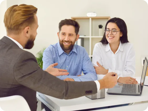 A mortgage broker sitting across from a smiling couple during a consultation, with a laptop on the desk, symbolizing financial planning and advice.