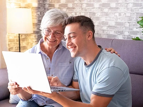 A happy young man and an older woman share a laptop on a couch, smiling as they use a guarantor calculator to explore home loan options.