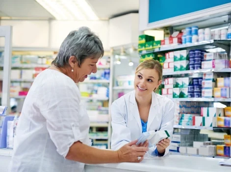 Pharmacist smiling and assisting a customer at a pharmacy counter, surrounded by shelves of medicines.