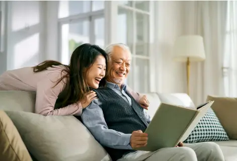 An elderly man and a young woman smile as they look through a photo album on a cozy couch in a well-lit living room.