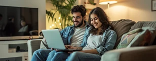 A man and woman in a blue shirt and white t-shirt sitting on a sofa looking at a laptop